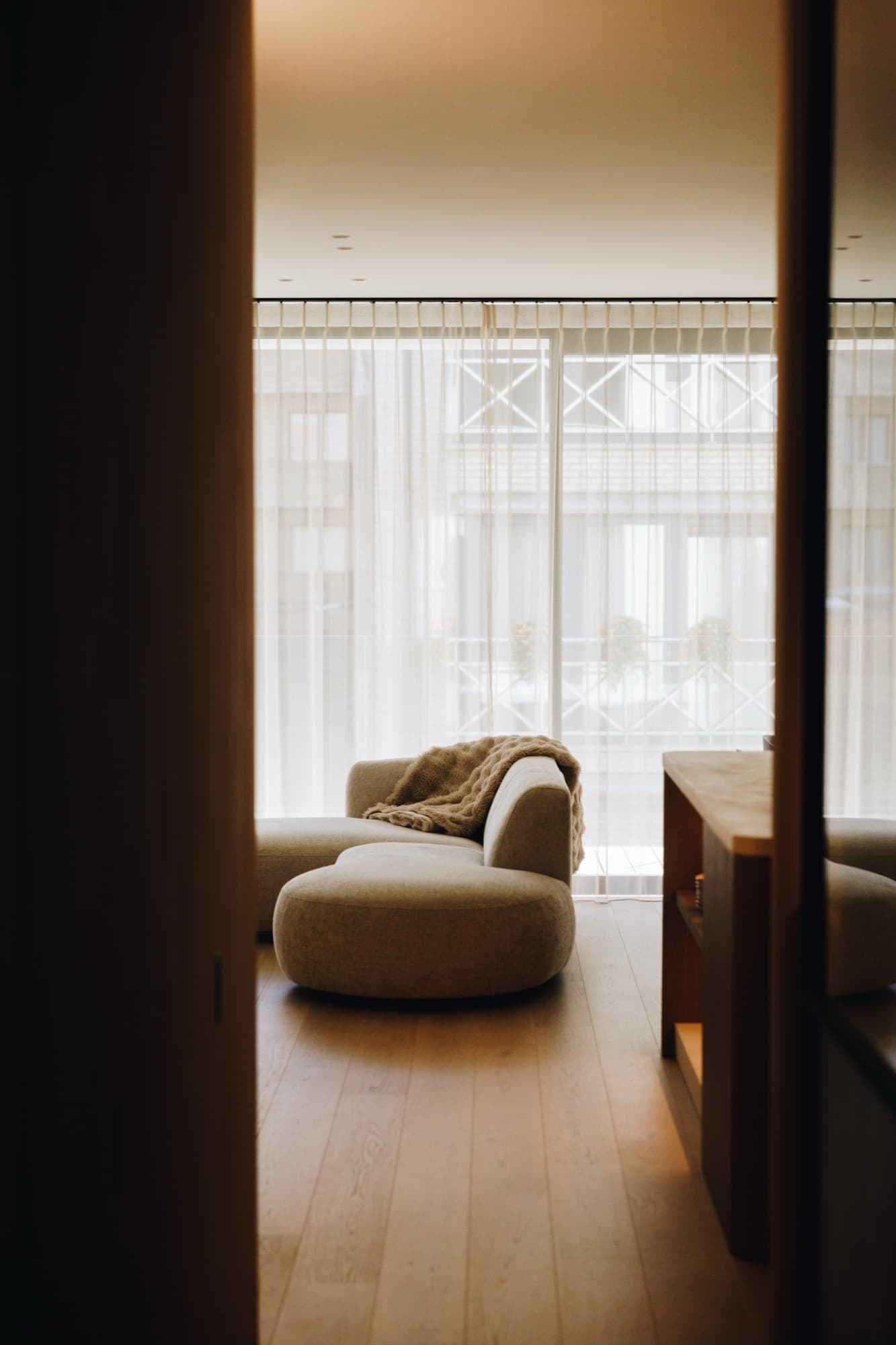 Doorkijk vanuit een gang naar een curved crème zetel met gebreide plaid, floor-to-ceiling vitrages en houten vloer.